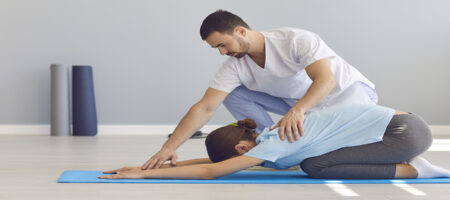 Side view the doctor helps the woman to do stretching and yoga exercises after the injury. Woman is lying on the mat face down with her legs raised and her arms outstretched. Concept of medical care.